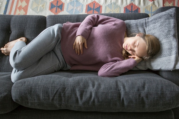 Kranke Frau, die zu Hause auf grauem Sofa, Hand auf Magen anhalten liegt