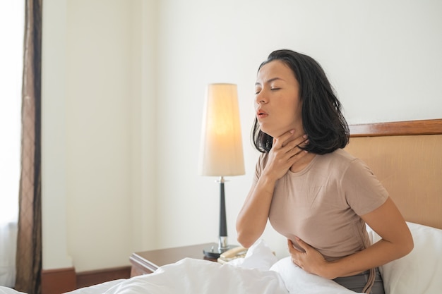Kranke Frau mit Husten- und Halsentzündung auf dem Bett, das sein Gesicht beim Husten bedeckt