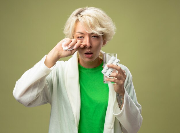 Kranke Frau mit kurzen Haaren, die sich unwohl fühlen, halten Glas Wasser und Blase mit Pillen, die ihre Nase mit Serviette abwischen, die über hellem Hintergrund steht