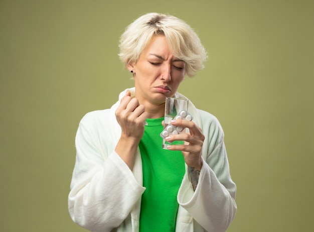 Kranke frau mit kurzen haaren, die sich unwohl fühlen, halten glas wasser und blase mit pillen, die über lichtwand stehen