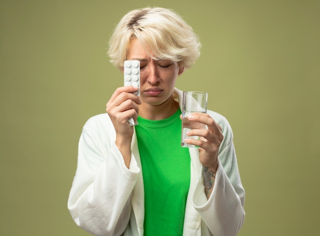 Kranke Frau mit kurzen Haaren, die sich unwohl fühlen, halten Glas Wasser und Blase mit Pillen mit geschlossenen Augen und traurigem Ausdruck, der über hellem Hintergrund steht