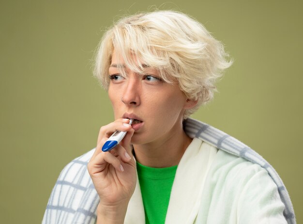 Kranke ungesunde Frau mit kurzen Haaren, die in eine Decke gewickelt sind, fühlen sich unwohl, wenn sie das Thermometer in den Mund stecken und an einer Grippe leiden, die über einer hellen Wand steht