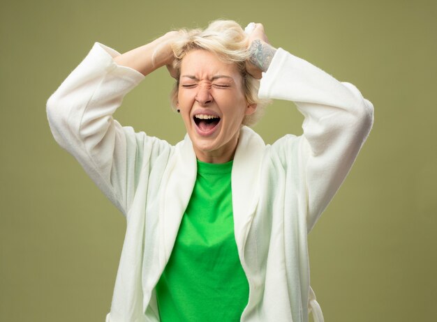 Kranke ungesunde Frau mit kurzen Haaren, die sich unwohl fühlen, wenn sie frustriert sind und ihr Haar über eine helle Wand ziehen