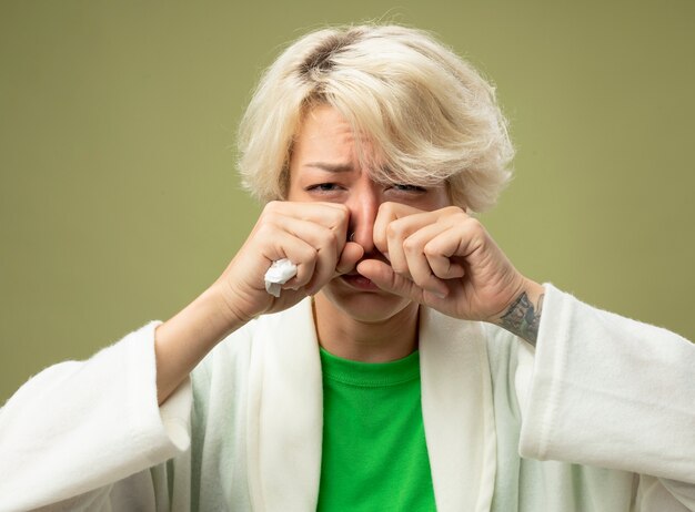 Kostenloses Foto kranke ungesunde frau mit kurzen haaren, die sich unwohl fühlen, wenn sie verärgert sind und ihre augen reiben, die an einer laufenden grippe-nase leiden, die über hellem hintergrund steht