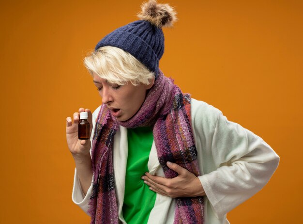 Kranke ungesunde Frau mit kurzen Haaren im warmen Schal und dem Unwohlsein, das Medizin hält, die unter Kälte leidet, die über orangeer Wand steht