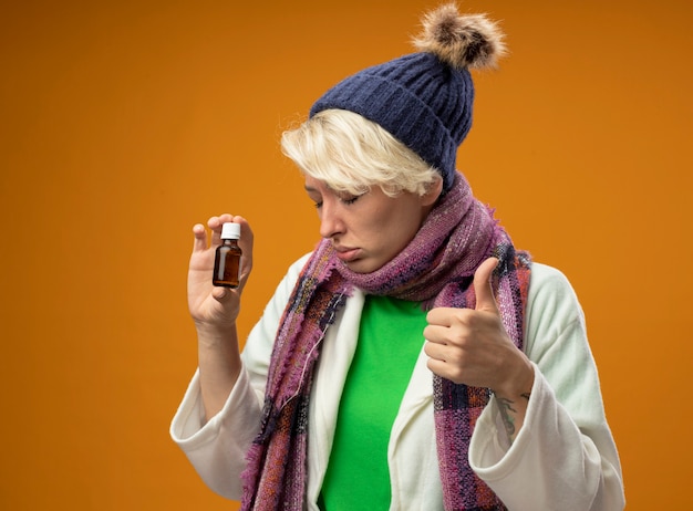 Kostenloses Foto kranke ungesunde frau mit kurzen haaren in warmem schal und hut, die medizinflasche hält, die daumen oben mit traurigem ausdruck über orange hintergrund steht