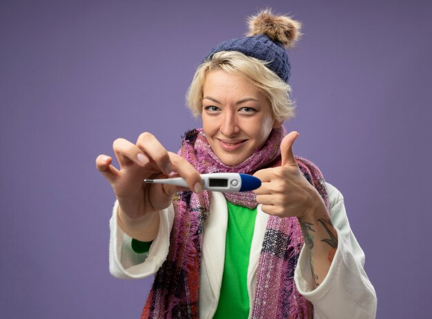 Kranke ungesunde Frau mit kurzen Haaren in warmem Schal und Hut, die sich besser fühlen, wenn sie das Thermometer lächelnd zeigt und Daumen hoch zeigt, die über lila Wand stehen