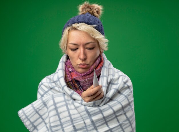 Kranke ungesunde Frau mit kurzen Haaren in warmem Schal und Hut, die sich unwohl fühlen, eingewickelt in Decke, die Thermometer hält, das über grünem Hintergrund steht