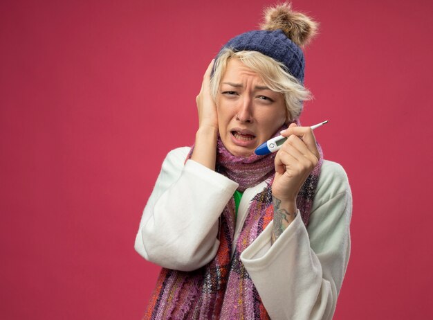 Kranke ungesunde Frau mit kurzen Haaren in warmem Schal und Hut, die sich unwohl fühlen, halten Thermometer baing in der Panik, die über rosa Wand steht