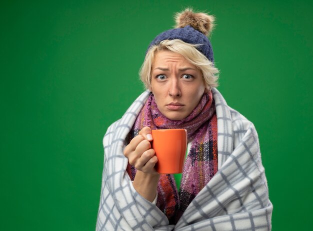 Kranke ungesunde Frau mit kurzen Haaren in warmem Schal und Hut fühlen sich unwohl eingewickelt in Decke, die Tasse Tee hält, die über grüner Wand steht