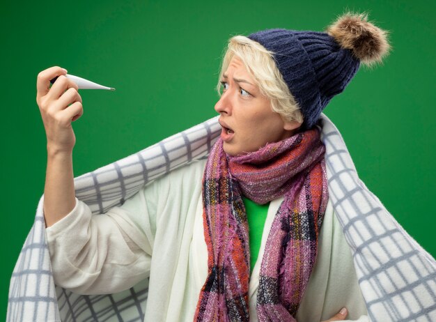 Kranke ungesunde Frau mit kurzen Haaren in warmem Schal und Hut fühlen sich unwohl eingewickelt in Decke gewickelt auf Thermometer, besorgt besorgt über grüner Wand stehend