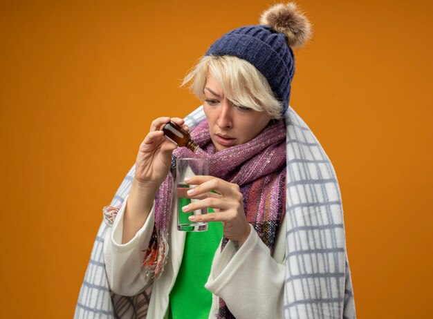 Kranke ungesunde Frau mit kurzen Haaren in warmem Schal und Hut gewickelt in Decke tropfende Medizin tropft in ein Glas, das über orange Hintergrund steht