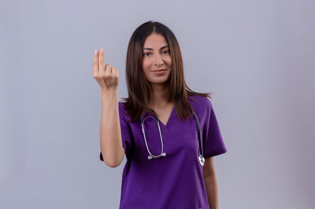 Krankenschwester der jungen Frau, die Uniform und mit Stethoskop trägt und zwei Finger zeigt, die freundliches Stehen lächeln