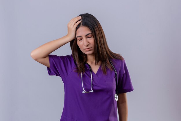 Krankenschwester der jungen Frau in der medizinischen Uniform und mit dem Stethoskop, das unwohl steht und mit der Hand auf Kopf steht, die unter starken Kopfschmerzen leidet