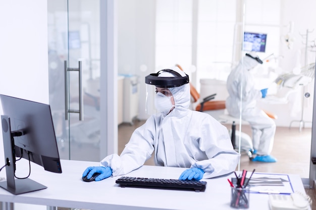Krankenschwester, die den Computer während Covid 19 verwendet, trägt einen PSA-Anzug als Sicherheitsvorkehrung. Medizinteam trägt Schutzausrüstung gegen Coronavirus-Pandemie in der zahnärztlichen Aufnahme als Sicherheitsvorkehrung.