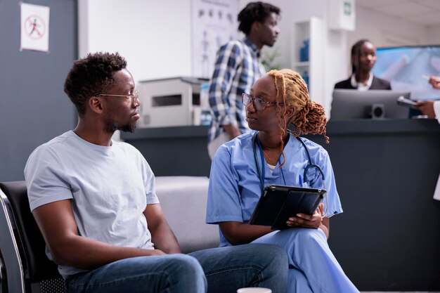 Krankenschwester, die sich mit dem Patienten berät, Notizen auf dem digitalen Tablet macht und im Wartezimmer Bericht schreibt. Arzthelferin im Gespräch mit Mann, Beratung einer Person mit Krankheit im Wartebereich.