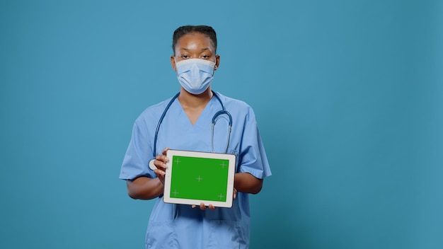 Krankenschwester, die Tablet mit horizontalem grünem Bildschirm hält und Gesichtsmaske gegen Coronavirus trägt. Facharzt mit modernem Gerät mit Chroma-Key, Mockup-Vorlage und isoliertem Hintergrund.