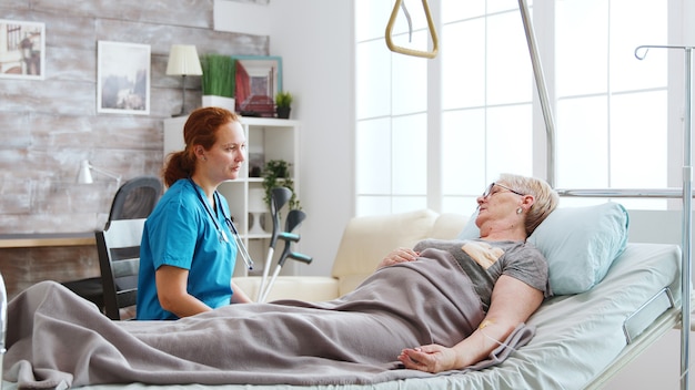 Krankenschwester im Altersheim im Gespräch mit einer alten Dame, die im Krankenhausbett liegt. Dahinter befinden sich große Fenster mit hellem Licht