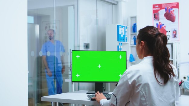 Krankenschwester in blauer Uniform betritt den Krankenhausschrank, während der Arzt einen Computer mit grünem Bildschirmmodell verwendet.