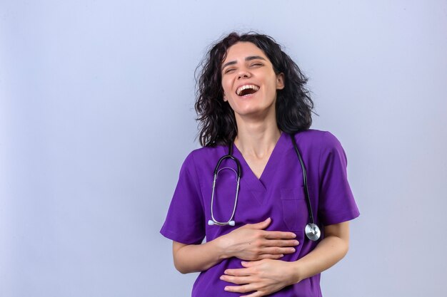 Krankenschwester in medizinischer Uniform und mit Stethoskop, das über guten Witz lacht und die Hände gekreuzt hält, die auf isoliertem Weiß stehen