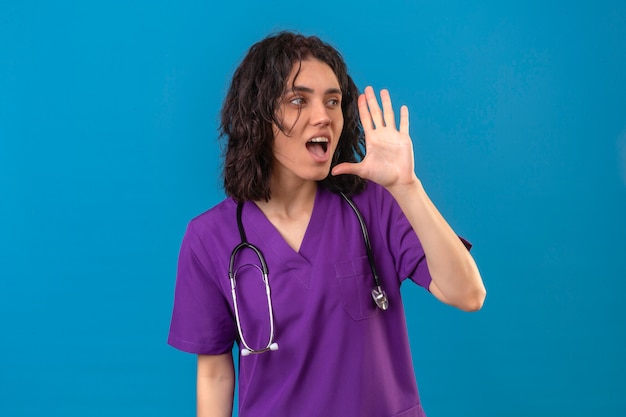 Krankenschwester in Uniform und Stethoskop schreit und ruft jemanden mit der Hand in der Nähe des Mundes auf isoliertem Blau