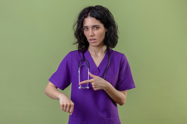 Krankenschwester in Uniform und Stethoskop zeigt auf ihre Hand und erinnert an die Zeit mit skeptischem Gesichtsausdruck auf einem isolierten Grün