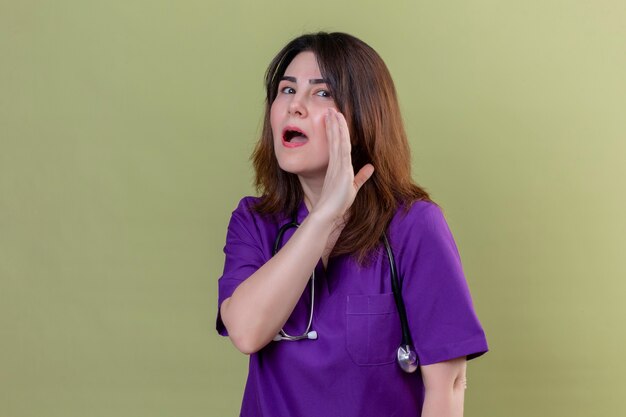 Krankenschwester mittleren Alters, die Uniform und mit Stethoskop mit einer Hand nahe dem Mund trägt, der ein Geheimnis erzählt, das über lokalisiertem grünem Hintergrund steht