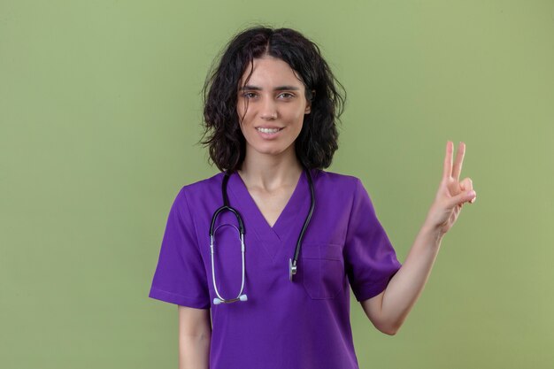 Krankenschwester trägt Uniform und Stethoskop lächelnd und zeigt Nummer zwei mit den Fingern oben auf isoliertem Grün