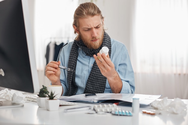 Kranker bärtiger männlicher Büroangestellter in blauem Hemd und Schal mit Brille konzentrierte sich auf das Lesen der Verschreibung von Pillen. Junger Manager mit Grippe, sitzt am Arbeitsplatz, umgeben von Drogen, Tabletten, Vitaminen