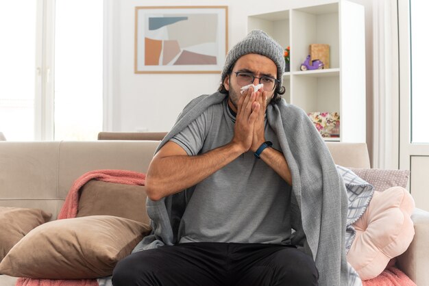 Kranker junger kaukasischer Mann in optischer Brille, eingehüllt in karierte Wintermütze, wischt sich die Nase mit Taschentuch ab und schaut auf die Kamera, die auf der Couch im Wohnzimmer sitzt