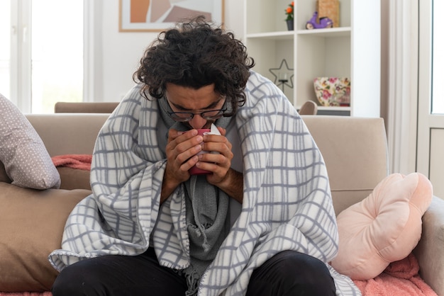 Kostenloses Foto kranker junger kaukasischer mann in optischer brille eingewickelt in plaid mit schal um den hals, der die tasse auf der couch im wohnzimmer hält und betrachtet