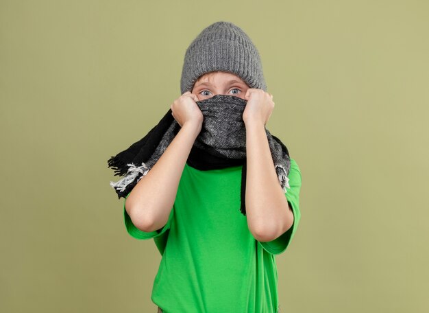 Kranker kleiner Junge, der grünes T-Shirt im warmen Schal und im Hut trägt, der mit Schal bedeckt sein Gesicht, das besorgt aussieht, über heller Wand stehend