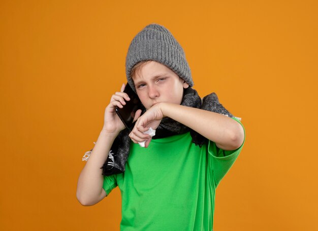 Kranker kleiner Junge, der grünes T-Shirt im warmen Schal und im Hut trägt und sich unwohl fühlt, wenn er auf Handy spricht, das Nase mit Papierserviette abwischt, die über orange Wand steht