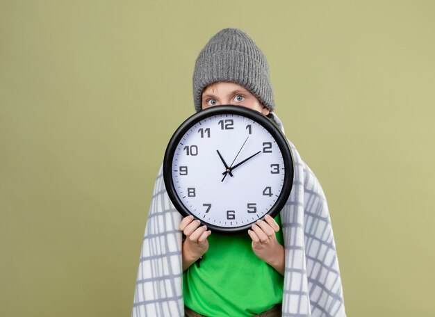 Kranker kleiner Junge, der grünes T-Shirt im warmen Schal und im Hut wickelt, der in Decke gewickelt hält, die Wanduhr hindendes Gesicht hinter der Uhr hält, die über heller Wand steht