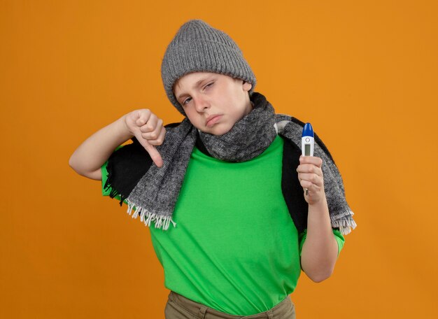 Kranker kleiner Junge, der grünes T-Shirt im warmen Schal und in der Mütze hält, die Thermometer halten, das sich unwohl krank und unglücklich fühlen, Daumen unten zeigend über orange Wand stehend