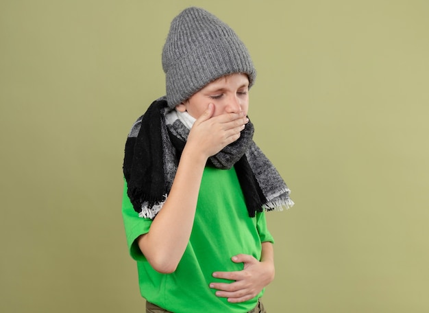 Kranker kleiner Junge, der grünes T-Shirt in warmem Schal und Hut hustet und sich unwohl fühlt, über heller Wand stehend