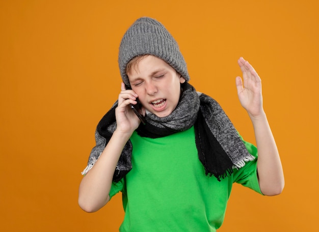 Kranker kleiner Junge, der grünes T-Shirt in warmem Schal und Hut trägt, die sich unwohl fühlen, auf Handy zu sprechen, unzufrieden mit genervtem Ausdruck, der über orange Wand steht