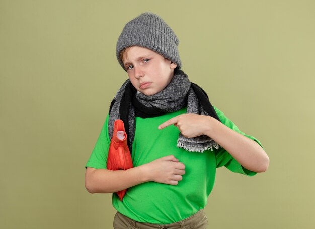 Kranker kleiner Junge, der grünes T-Shirt in warmem Schal und Hut trägt, die sich unwohl fühlen, Wasserflasche zu halten, um warm zu halten, mit dem Finger darauf zeigend über heller Wand stehend