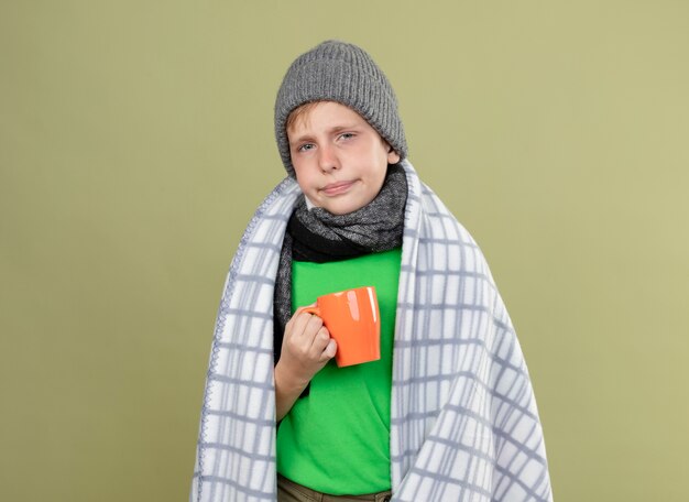 Kranker kleiner Junge, der grünes T-Shirt in warmem Schal und Hut wickelt, die in Decke gewickelt halten Tasse des heißen Tees halten unglücklich und krank stehen über heller Wand