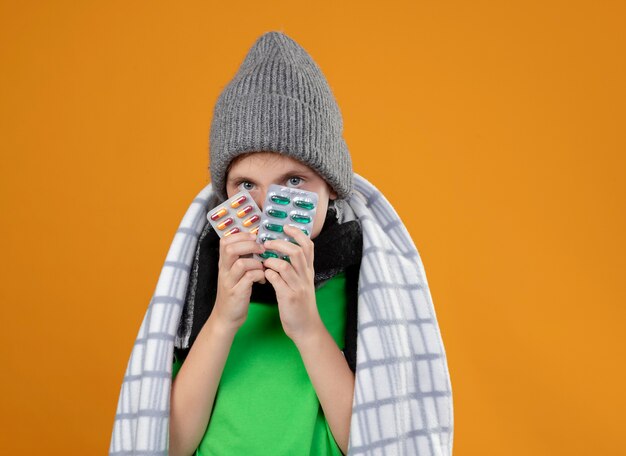 Kranker kleiner Junge, der warmen Hut und Schal trägt, die in die Decke gewickelt werden, die unter Fieber leidet, das Pillen hält, die sich unwohl fühlen, die über der orangefarbenen Wand stehen