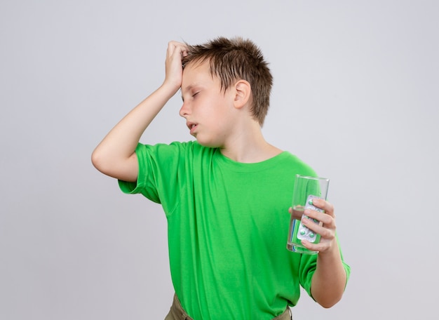 Kranker kleiner Junge im grünen T-Shirt, der sich unwohl fühlt und ein Glas Wasser und Pillen hält, die seinen Kopf berühren, der unter Kälte leidet, die über weißer Wand steht