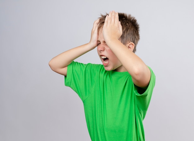 Kranker kleiner Junge im grünen T-Shirt, der sich unwohl fühlt und starke Kopfschmerzen hat, die Kopf berühren, der über weißer Wand steht