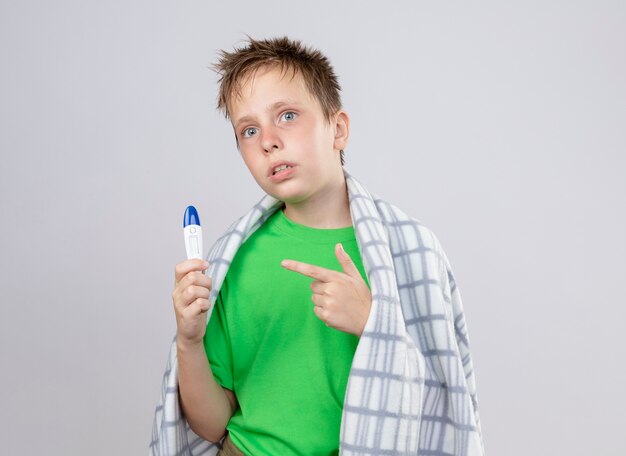 Kranker kleiner Junge im grünen T-Shirt, eingewickelt in Decke, die sich unwohl hält, Thermometer haltend, das mit Zeigefinger darauf zeigt, der über weißer Wand steht