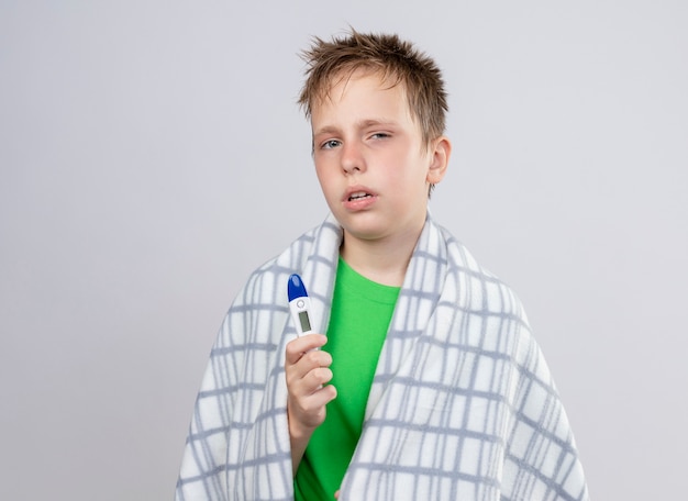 Kranker kleiner Junge im grünen T-Shirt, eingewickelt in Decke, die sich unwohl hält, Thermometer haltend über weißer Wand