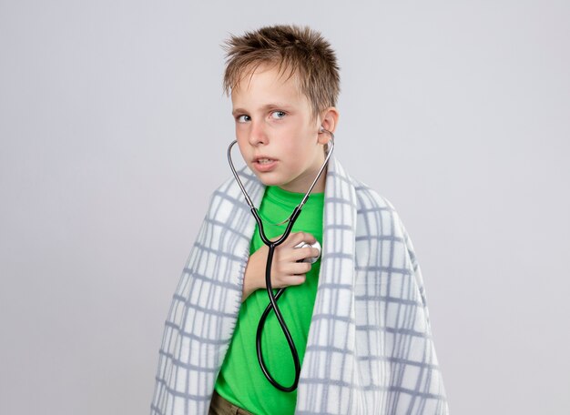 Kranker kleiner Junge im grünen T-Shirt, eingewickelt in die Decke, die sich mit Stethoskop um seinen Hals unwohl fühlt und lauscht, wie der Herzschlag verwirrt ist, der über weißer Wand steht