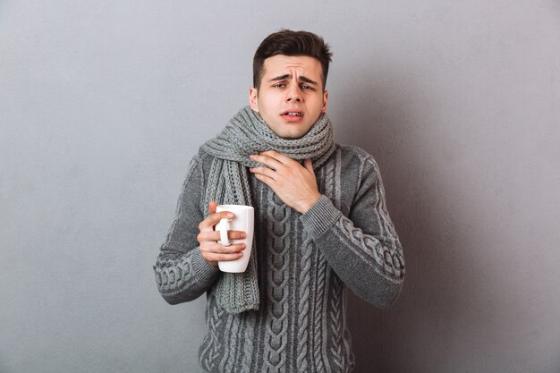 Kranker Mann in der Strickjacke und in Schal, die Halsschmerzen beim Halten der Tasse Tee haben