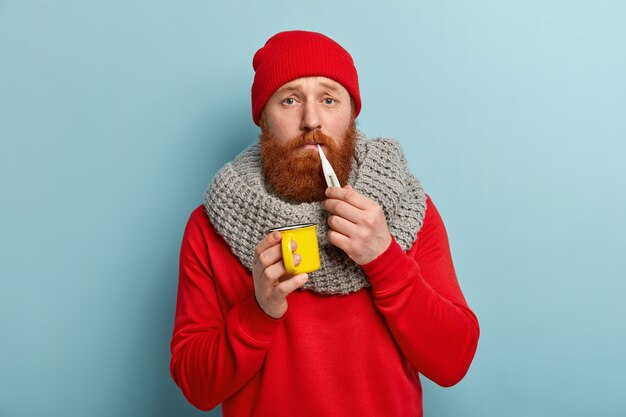 Kranker Mann in warmer Kleidung mit Thermometer und Tasse