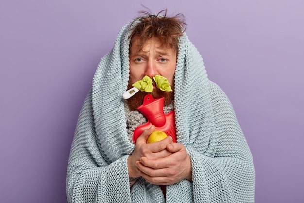 Kranker Mann in warmer Kleidung mit Thermometer und Wassersack