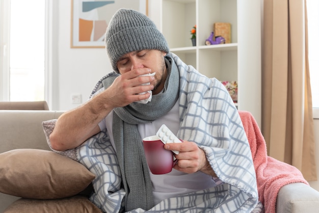 Kranker Mann mit Schal um den Hals, der eine Wintermütze trägt, wischt sich die Nase ab und hält eine Tasse auf der Couch im Wohnzimmer