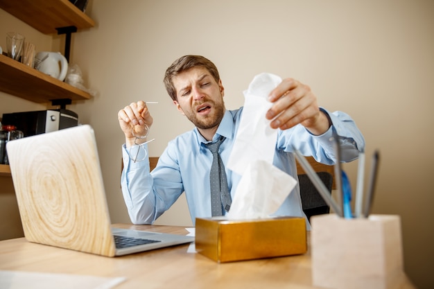 Kranker Mann mit Taschentuch Niesen putzend Nase während der Arbeit im Büro, Geschäftsmann erkältet, saisonale Grippe. Influenza-Pandemie, Krankheitsvorbeugung, Klimaanlage im Büro verursachen Krankheit